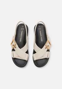 Sandales à bride croisée beige avec une boucle dorée et une semelle noire. La partie supérieure présente une texture lisse et mate, et la semelle intérieure est imprimée avec les détails de la marque.