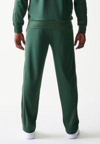 Πράσινο παντελόνι φόρμας από πολυεστέρα με στενή γραμμή, λευκές πλαϊνές ρίγες και ελαστικές μανσέτες, συνδυασμένο με λευκά αθλητικά παπούτσια.