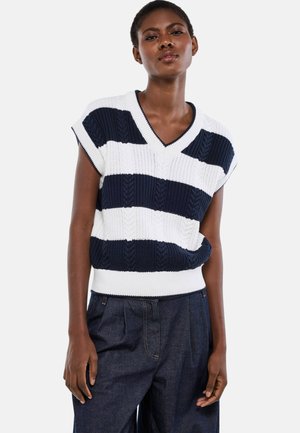 Femme portant un gilet en tricot sans manches à rayures bleu marine et blanches avec un col en V et un pantalon large foncé, debout devant un fond uni.