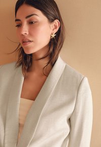 Lichtgrijze blazer met een overslagontwerp en een gladde textuur, gecombineerd met een beige top en gouden oorbellen in de vorm van sterren, tegen een neutrale achtergrond.