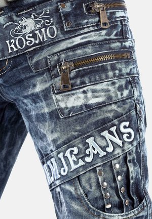 Denim cargo pants in dark blue with a faded pattern. Features include multiple zippered pockets, embroidered logo, and decorative studs.