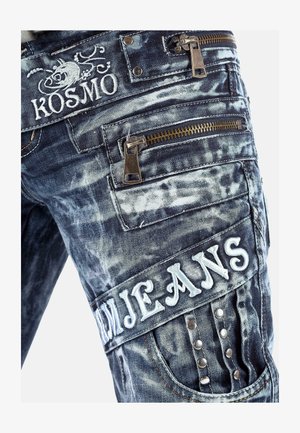 Pantaloni cargo in denim di colore blu scuro con un motivo sbiadito. Caratteristiche includono molteplici tasche con zip, logo ricamato e borchie decorative.
