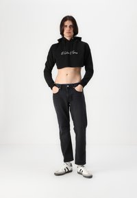 Black cropped hoodie with embroidery at the chest, paired with dark jeans and white sneakers featuring black stripes. Plain background.