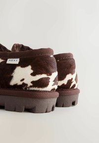 Scarpe slip-on a stampa mucca marrone e bianco con tomaia in pelliccia testurizzata, bordo in tessuto marrone a contrasto e suola in gomma resistente.
