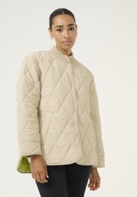 Beige gewatteerde jas met ruitpatroon, geribde kraag en drukknoopsluiting. Voering van contrasterende groene stof bij de manchetten.