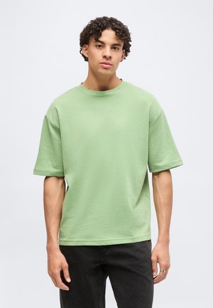 Jonge man met krullend haar die een lichtgroen oversized T-shirt en zwarte broek draagt, staand tegen een effen achtergrond.