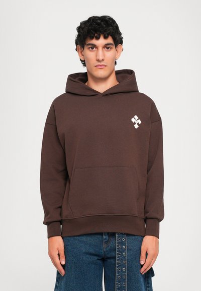 Giovane uomo con capelli corti e ricci che indossa un hoodie marrone con un piccolo logo bianco sul petto e jeans blu scuro su uno sfondo uniforme.