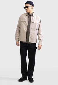 Giacca beige leggera con chiusura a bottoni sul davanti, due tasche sul petto, indossata sopra una camicia a quadri marrone, abbinata a pantaloni neri e calzature.