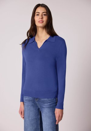 Donna che indossa un maglione blu a collo a V con maniche lunghe e un colletto, abbinato a jeans azzurri, in piedi davanti a uno sfondo chiaro e uniforme.