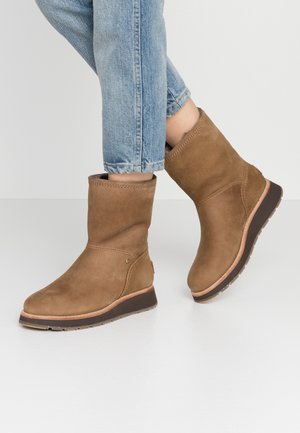 Brązowe skórzane botki na płaskiej, ciemnej podeszwie noszone z jasnoniebieskimi podwiniętymi jeansami na białej podłodze.