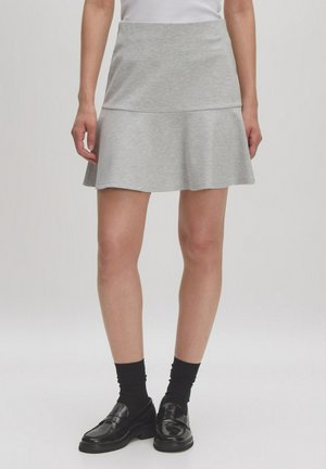 Grauer Baumwollrock mit einem ausgestellten Saum, ausgestattet mit einem figurbetonten Bund und geschichtetem Design. Trägt schwarze Schuhe und Socken.
