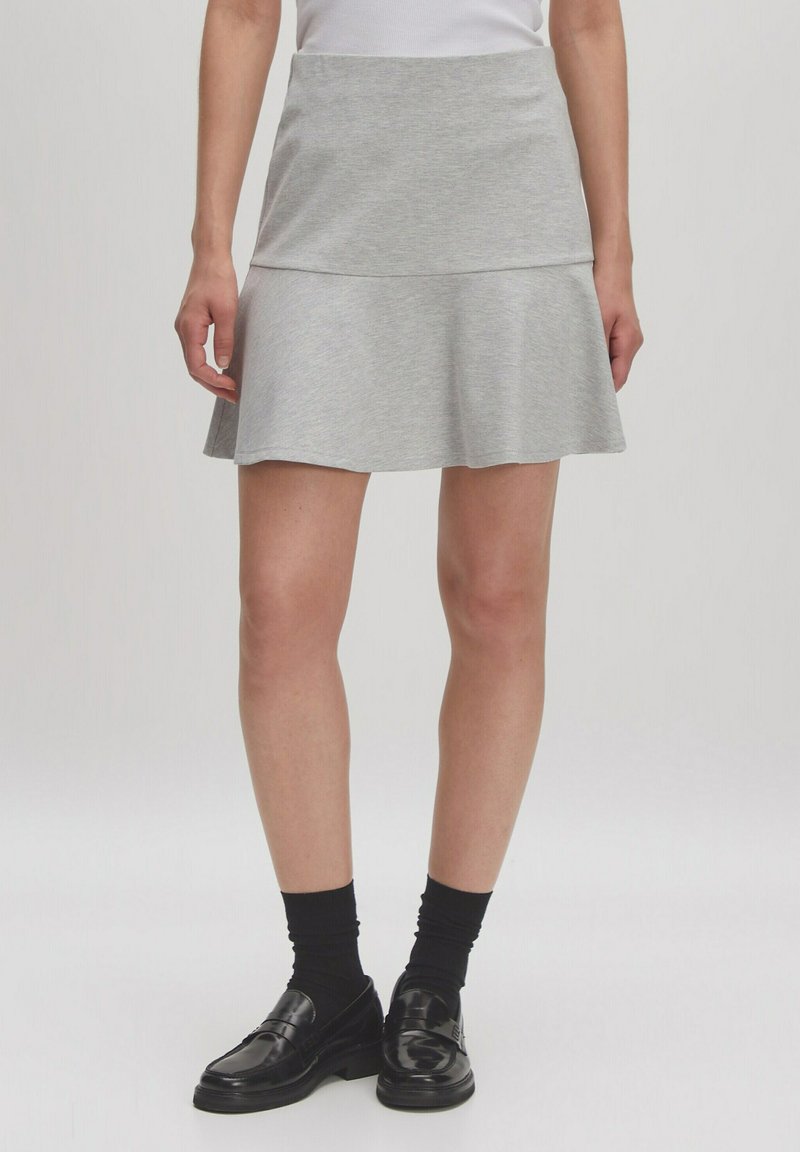 Grauer Baumwollrock mit einem ausgestellten Saum, ausgestattet mit einem figurbetonten Bund und geschichtetem Design. Trägt schwarze Schuhe und Socken.