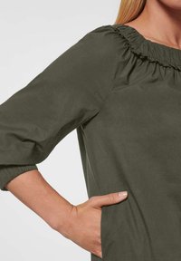 Olivengrünes Kleid mit gerafftem Ausschnitt, 3/4-Ärmeln und elastischen Manschetten. Verfügt über seitliche Taschen für zusätzliche Funktionalität. Glatte Textur.