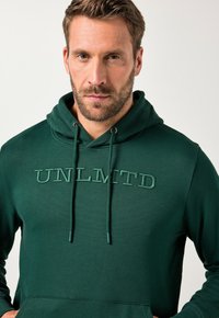 Felpa con cappuccio verde scuro, con tasca a marsupio, cappuccio regolabile con cordino e testo ricamato "UNLMTD" sul petto con filo abbinato.