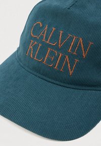 Sinine lõngakanga müts, millel on rusti värvi tikand "CALVIN KLEIN" logo. Omab kõverat ääre ja tekstureeritud kangast.