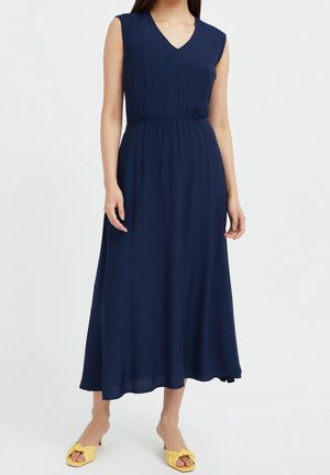 Kobieta ubrana w granatową sukienkę midi bez rękawów z dekoltem w kształcie litery V oraz żółte sandały na obcasie, stojąca na tle białej ściany.