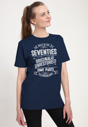 Kvinde med en lang hestehale iført en mørkeblå T-shirt med hvid tekst, der lyder: "Bygget i 70'erne, Original og Urestaureret, Nogle dele stadig i funktionsdygtig stand."