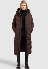 Brun pufferjakke med hætte, med et langt, quiltet design og sidelommer. Bæres over en sort turtleneck og sorte bukser.