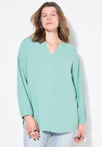 Blusa verde chiaro a V con maniche lunghe, texture setosa, vestibilità rilassata e polsini elasticizzati. Indossata su jeans blu. Design semplice.