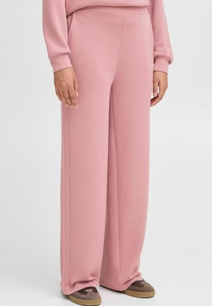 Wijde broek in zacht roze stof, met een gladde textuur en een elastische tailleband. Draagt bij bruine schoenen, wat zorgt voor een relaxte pasvorm.