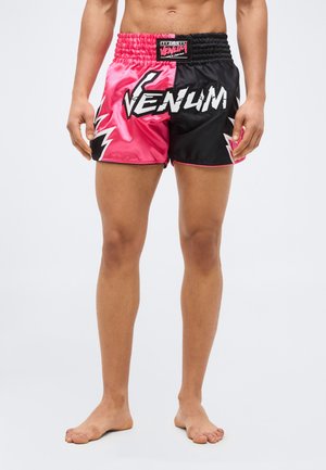 Bărbat purtând pantaloni scurți de marcă Venum, roz și negri, cu grafice cu fulgere, stând desculț pe un fundal simplu.