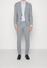 Traje gris de corte slim, combinado con una camisa azul con estampado. Los accesorios incluyen zapatos negros con detalle de hebilla dorada.