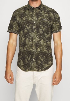 Mies, jolla on päällään lyhythihainen, napitettava paita, jossa on tummanvihreä ja musta saniaiskuvio, yhdistetty vaaleisiin housuihin, seisoo valkoisen taustan edessä.