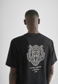 Camiseta de algodón negra con un gráfico de tigre blanco y el nombre de la marca en la parte posterior. Manga corta y un corte holgado. Textura suave.