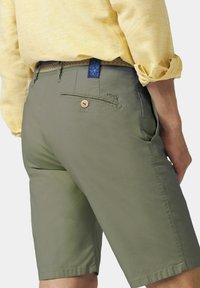 Shorts vert olive avec une finition lisse, présentant une poche arrière avec un bouton en bois et un logo brodé, associés à une chemise jaune clair.