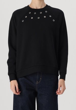 Personne portant une chemise noire à manches longues avec des œillets argentés près du col et un pantalon en jean bleu foncé.