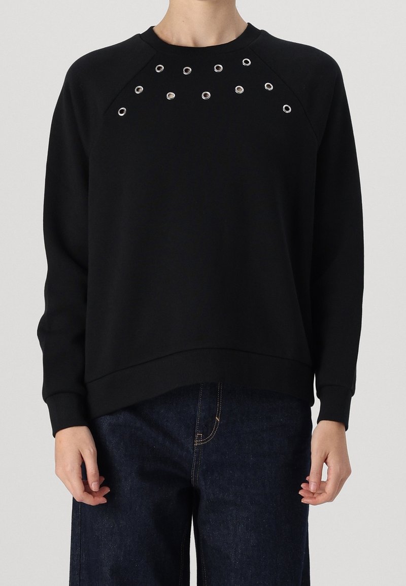 Personne portant une chemise noire à manches longues avec des œillets argentés près du col et un pantalon en jean bleu foncé.