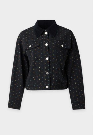 Femme portant une veste courte noire à motif, une chemise rayée bleue, une jupe marine avec un ourlet en dentelle et des chaussures Mary Jane bordeaux, debout avec la main sur la hanche.