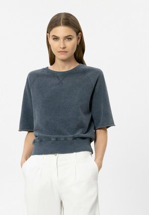 Frau mit glattem braunem Haar, die ein ausgewaschenes blaues Kurzarm-Sweatshirt und weiße hochtaillierte Hosen trägt, mit den Händen in den Taschen.