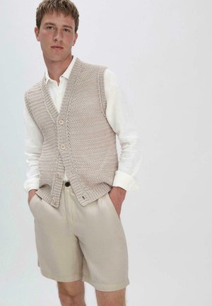 Beige gestrickte Weste mit V-Ausschnitt, die ein strukturiertes Muster und drei Knöpfe aufweist. Über einem weißen Langarmshirt und beigen Shorts getragen.