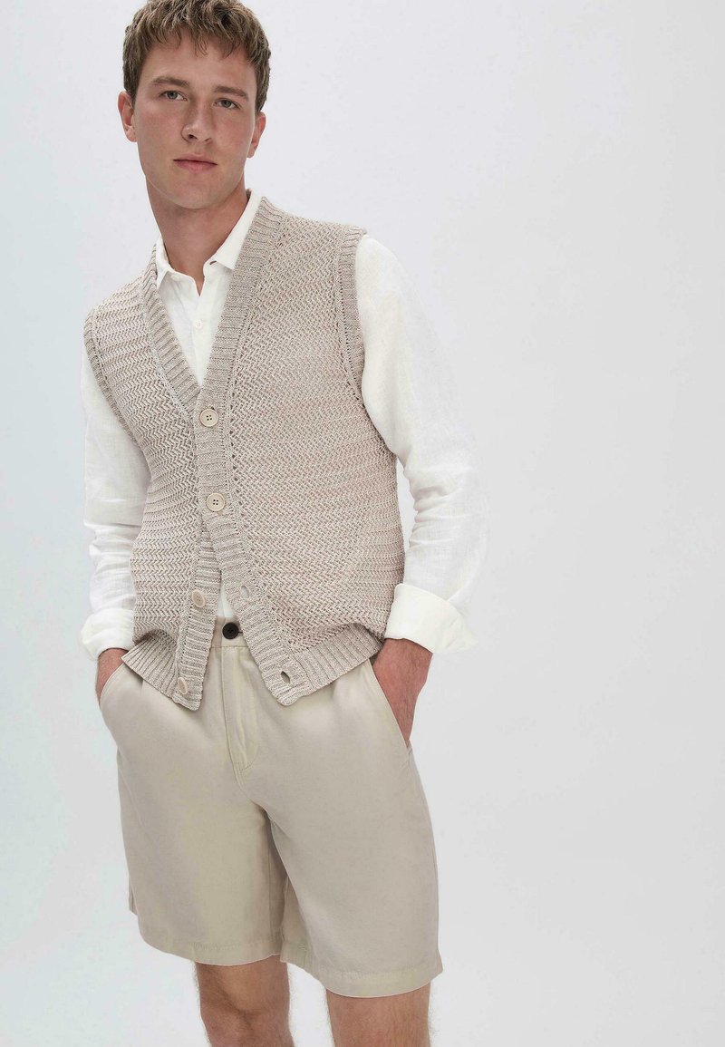 Beige gebreide vest met een V-hals, voorzien van een gestructureerd patroon en drie knopen. gedragen over een wit langesleeve shirt en beige shorts.