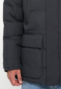 Dark padded jacket featuring a matte texture, large front pocket, and adjustable cuffs, paired with light blue denim jeans.