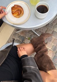 Bota de ante marrón hasta la rodilla al lado de una falda negra y una mano sobre la rodilla; plato con un pastel trenzado y taza blanca de café sobre una mesa de plata.