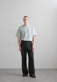 T-shirt bleu clair à manches courtes associé à un pantalon noir plissé taille haute. Le mannequin se tient sur un sol gris devant un arrière-plan neutre.