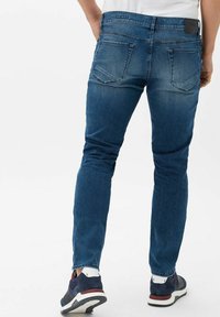 Blue denim jeans with a slim fit, five pockets, and a black waistband label. Worn with navy sneakers featuring white accents.