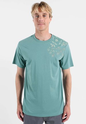 Homme blond portant un t-shirt vert avec un motif floral beige subtil sur l'épaule droite, debout devant un fond blanc uni.