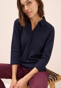 Tmavě modré polo tričko s dlouhým rukávem a límečkem, třemi knoflíky a měkkou texturou, doplněné o mahagonové kalhoty. Jednoduchý design, minimální akcenty.