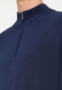 Granatowy sweter z dzianiny z wysokim prążkowanym kołnierzem, pół-zamkiem oraz gładką teksturą. Skup się na detalach kołnierza i zamka.