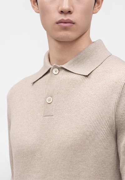 Jeune personne portant un polo beige en maille avec un col et deux boutons, visible de la poitrine vers le haut sur un fond uni.