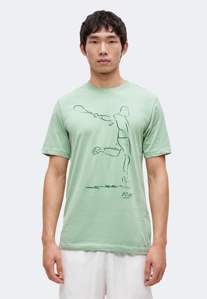 Jeune homme portant un t-shirt vert clair avec un dessin au trait d’un joueur de tennis frappant une balle à l’avant, associé à un short blanc.