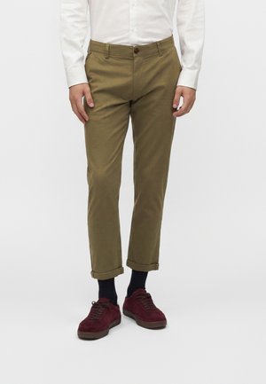 Man die een opgerolde olijfgroene broek draagt, zwarte sokken, bordeauxrode sneakers en een wit overhemd met knopen tegen een effen witte achtergrond.