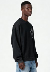 Jeune homme de profil portant un sweat-shirt noir avec une croix blanche et le texte "KARMA KILL", associé à un jean bleu clair.