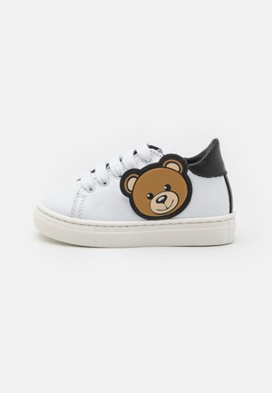 Scarpe da ginnastica bianche in materiale sintetico, con retro nero, suola rotonda in gomma e una grande toppa a forma di faccia di orso bruno sul lato.