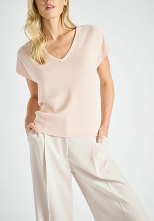 Vrouw die een lichtroze V-hals top met korte mouwen en loszittende crèmekleurige broek draagt met handen in de zakken tegen een witte achtergrond.