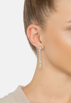 Long drop earring with multiple clear gems and a larger yellow gemstone at the bottom, worn on a woman's ear with hair pulled back.