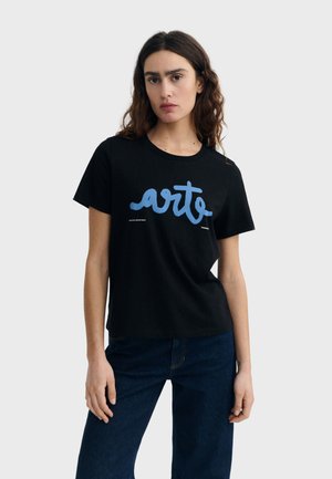 Kobieta o falujących brązowych włosach, ubrana w czarną koszulkę z niebieskim napisem „arte” oraz ciemnoniebieskie dżinsy, stojąca na jednolitym tle.